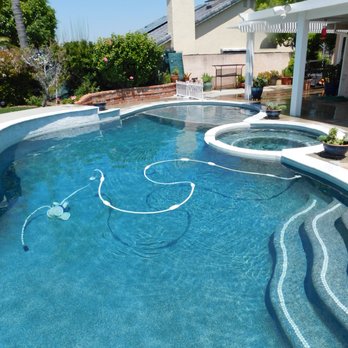 Poolscape Unlimited - Love it - A backyard scene featuring a swimming pool with a hot tub attached, filled with clear water. The pool has built-in steps on the right side and is bordered by a white concrete edge. The surrounding patio area includes potted plants, outdoor furniture, and a shaded section with a pergola. In the background, there are trees, shrubs, and a house with a chimney, under a clear blue sky. - Pool & Hot Tub Service Near Me - Lakeside, California