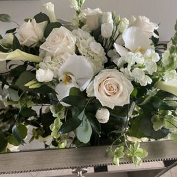 A floral arrangement with white roses, orchids, and other white flowers, complemented by green leaves and stems, is displayed in a glass vase on a table. The bouquet includes various flower types and lush foliage, creating a full and elegant presentation suitable for a formal setting or special occasion.