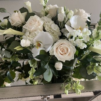Mangel - A floral arrangement with white roses, orchids, and other white flowers, complemented by green leaves and stems, is displayed in a glass vase on a table. The bouquet includes various flower types and lush foliage, creating a full and elegant presentation suitable for a formal setting or special occasion. - Florists Near Me - Chicago, Illinois