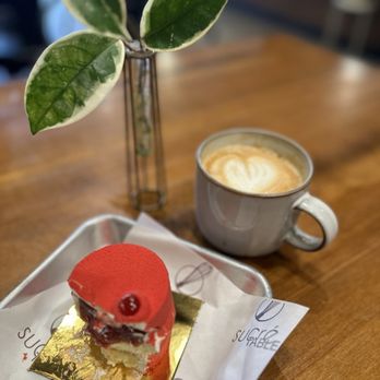Sucre Table - The image shows a small, decorated cake with a red glaze and a single berry on top, placed on a gold-colored base and a branded paper wrapper. Next to it is a gray ceramic mug filled with a latte, featuring a heart-shaped latte art design. In the background, a potted plant with variegated green and white leaves is positioned on the table. The setting appears to be a cafe with a wooden table surface and a blurred interior background. - Bakeries Near Me - Tampa, Florida