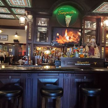 Tempesta's - a bar with bar stools - A bar with a dark wood counter, three bar stools, and a display of liquor bottles is shown inside Tempesta's restaurant, with decorative lighting and a television screen. - American Near Me - Keene, New Hampshire