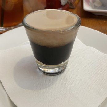 Irish Times - China White - A small, clear glass holds a layered coffee drink with dark coffee at the bottom and a light, creamy foam or milk layer on top. The glass is set on a white napkin, which rests on a plate. In the background, there are various condiments, bottles, and dishes on a wooden table, indicating a dining setting. - Pubs Near Me - Brookfield, Illinois