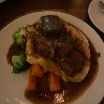 Moro's Kitchen - A plate contains beef stew with a piece of beef topped with gravy, served over mashed potatoes. The dish includes cooked carrots and broccoli on the side, with gravy surrounding the food. The presentation is on a white round plate, set on a dark wooden table. - Italian Near Me - Skaneateles, New York