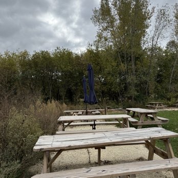 Aspen Sky - Multiple wooden picnic tables with attached benches are arranged outdoors on a gravel surface, with some closed navy blue umbrellas positioned behind them. The area is bordered by bushes and trees, with a cloudy sky overhead, suggesting an outdoor setting suitable for gatherings or dining. - Wineries Near Me - Slinger, Wisconsin