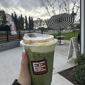 Febe Coffee - Banana pudding matcha - A person with manicured nails holds a clear plastic cup filled with a green matcha drink topped with whipped cream and a crumbly topping, from Coffee Bean & Tea Leaf. The outdoor setting includes a patio with a black-and-white striped umbrella, a small table, and a signboard. The background features leafless trees, a cloudy sky, and a road with cars, indicating a cool or overcast day. - Coffee & Tea Near Me - Irvine, California
