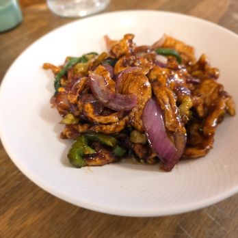 NiHao - Stir Fried Chicken with Mixed Peppers - The image shows a white, textured bowl filled with stir-fried chicken pieces, red onion slices, and green peppers, all coated in a glossy, dark sauce. The bowl is on a wooden table, with part of another dish visible on the left and some glassware in the background. The dish appears to be a Chinese-style stir-fry, likely served at a restaurant. - Chinese Near Me - Baltimore, Maryland