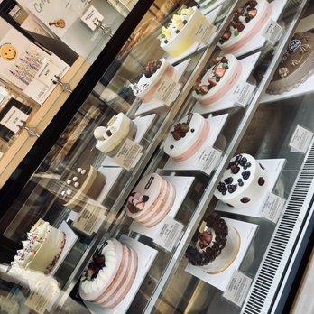 Tous Les Jours - Cakes and treats - A glass display case contains multiple decorated cakes arranged in rows, featuring various flavors, toppings, and designs. The cakes include fruit toppings, cream decorations, and different color schemes, indicating a selection of bakery or cake shop offerings. Labels with descriptions and prices are visible next to each cake, and the background shows part of the shop interior with signage and packaging. - Coffee & Tea Near Me - Midvale, Utah