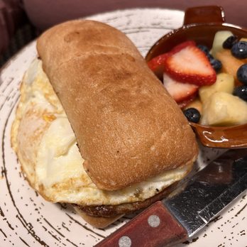 Honeyfield Restaurant - Sausage egg and cheese on a ciabatta roll - A breakfast plate includes a sandwich with a fried egg on a toasted ciabatta roll, placed on a decorative plate. Next to it is a small brown bowl filled with mixed fresh fruit, such as strawberries, blueberries, and banana slices. A serrated knife with a wooden handle rests on the plate, and part of a person's hand is visible in the background. - Breakfast & Brunch Near Me - Lockport, Illinois