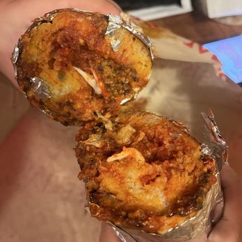 Massi's - Meatball Sub - A close-up image shows a partially eaten meatball sub sandwich wrapped in foil, with visible meatballs, tomato sauce, and melted cheese inside. The sandwich is held in a person's hand, with a blurred background that appears to be a wooden surface and some packaging. - Sandwiches Near Me - Astoria, New York