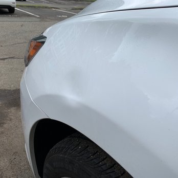 Mayhem Autoworks - The image depicts a white car with a noticeable dent on the front fender above the wheel. The vehicle is parked on a street with a paved surface, and other cars are visible in the background. The photo captures part of the car's front side, including the headlight and tire, with a focus on the damage to the fender. - Auto Customization Near Me - Wallingford, Connecticut