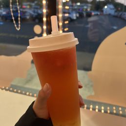 A hand is holding a large cup of bubble tea with a straw, positioned in front of a window decorated with string lights. Outside the window, a parking lot with multiple cars and a cloudy sky are visible during dusk. The interior features decorative string lights, creating a cozy ambiance.