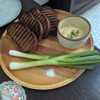 Sudova * Eastern European Kitchen - Khlib & Sil - A wooden plate holds grilled bread slices, green onions, and a bowl of creamy dip garnished with chives, with a salt pile nearby. - Modern European Near Me - Cincinnati, Ohio