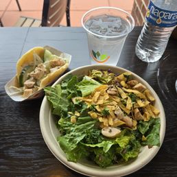 A salad with greens, chicken, and tortilla strips, a taco with chicken and vegetables, a bottle of Niagara water, and a cup of water on a dark wooden table.