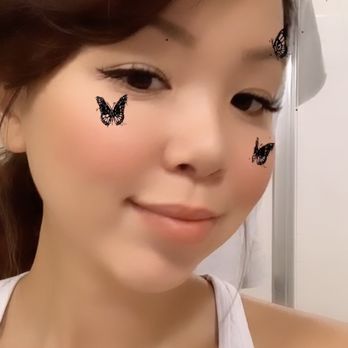 Mint Cloud TBP - Natural Lashes Extensions after a few days (wearing some liquid eyeliner here) - A woman with brown hair styled to one side, wearing makeup with eyeliner and blush, is taking a selfie. Butterfly stickers are placed on her cheeks and near her forehead. She is wearing a white sleeveless top, and the background appears to be indoors with a neutral wall and a metallic object visible. - Eyelash Service Near Me - Superior, Colorado