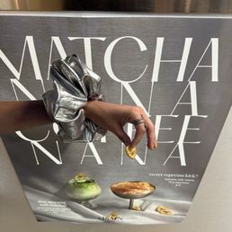 A hand with rings and a metallic silver scrunchie is holding a banana slice in front of a cafe menu poster. The poster advertises matcha-based drinks and desserts, with images of a matcha drink and a dessert on a white surface. The background includes part of a counter and a person wearing a striped shirt.