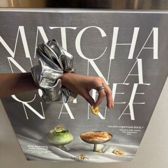 Cafe MSMN - A hand with rings and a metallic silver scrunchie is holding a banana slice in front of a cafe menu poster. The poster advertises matcha-based drinks and desserts, with images of a matcha drink and a dessert on a white surface. The background includes part of a counter and a person wearing a striped shirt. - Coffee & Tea Near Me - Brooklyn, New York