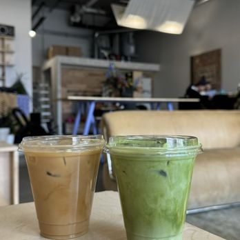 Drip Collective - Mannys matcha - Two iced beverages, one coffee and one matcha, are placed on a wooden table inside a coffee shop with modern decor and seating. - Coffee & Tea Near Me - Chicago, Illinois