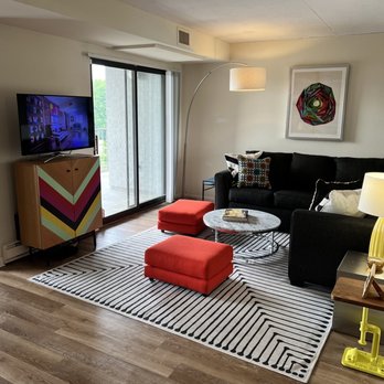 Concierge - Carlisle living room, bright windows sliding door to balcony - Living room with a black sofa adorned with patterned pillows, a round marble coffee table, and two red ottomans on a black-and-white striped rug. A sliding glass door opens to a balcony, allowing natural light. A colorful geometric artwork hangs on the wall, and a yellow table lamp is on a side table. A TV is positioned on a colorful cabinet near the sliding door. - Apartments Near Me - Rocky Hill, Connecticut