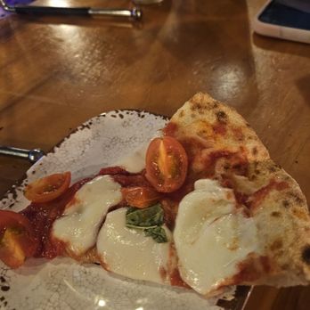 The Station - Yummmm - A single slice of Margherita pizza featuring melted mozzarella cheese, cherry tomato halves, and basil leaves is placed on a decorative ceramic plate with a crackle glaze pattern. The pizza has a slightly charred crust and is served on a wooden table in a dimly lit setting, with a glass, a smartphone, and other items visible in the background. - Pizza Near Me - Clermont, Florida