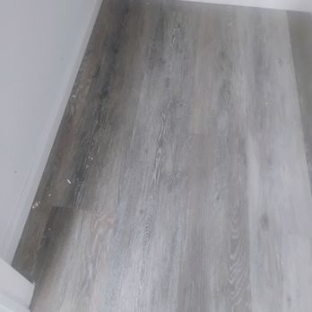 Clean Champs - A photograph captures a corner of an interior room featuring light gray wooden flooring with visible wood grain patterns. The walls are painted white, and there is a white baseboard along the bottom edge of the wall. Some dust and small debris are scattered on the floor, indicating recent work or cleaning activity. - Home Cleaning Near Me