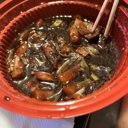 A red bowl holds a dark, thick sauce with pieces of meat and vegetables, possibly onions and green onions, in a savory Asian dish. The dish appears to be a stir-fry or braised preparation, garnished with sesame seeds. Two chopsticks are inserted into the bowl, and the bowl is placed on a white napkin on a dark surface.
