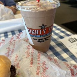 Blue Bell - A chocolate Blue Bell milkshake in a clear plastic cup with a domed lid and a red straw is positioned on a checkered table at Mighty Fine Burgers. Part of a burger wrapped in branded paper is visible in the foreground, and the background shows a casual restaurant interior with shelves and a person seated nearby.