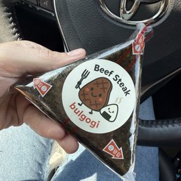 Bulgogi  Soy Sauce Bulgogi Beef - A person holds a triangular onigiri package labeled 'Beef Steak bulgogi' inside a car, with the Toyota logo visible on the steering wheel.