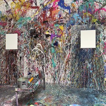 The Splatter Studio - Paint canvas - A room with a wall covered in multicolored paint splatters and drips, creating an abstract art background. Two blank white canvases are mounted on the wall. A table with painting tools, including a bucket, roller, and brushes, is positioned in front of the wall. The floor is also splattered with paint, indicating active use of the space for creative painting activities. - Team Building Activities Near Me - Atlanta, Georgia
