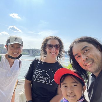 DC Design Tours - Five people, including a child, are taking a selfie outdoors near a river with a bridge and cityscape visible in the background. The group includes three adults and one child, all smiling at the camera. The sky is clear with some clouds, and the scene appears to be during daytime. One person is wearing a DC Design Tours t-shirt, indicating they may be participating in a tour or outdoor activity. - Architectural Tours Near Me