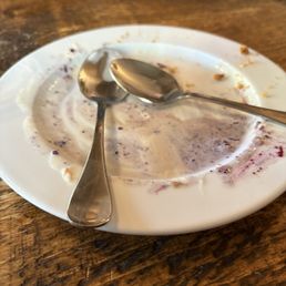 This was the remains of the blueberry pie - A white plate with leftover blueberry pie crumbs and sauce, accompanied by two metal spoons, on a wooden table surface.