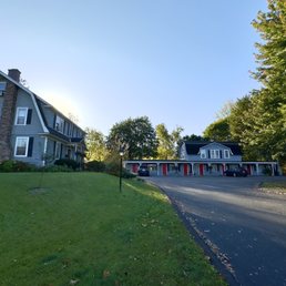 A large house with multiple windows and a chimney is situated on a grassy lawn, with a paved driveway leading to a garage with red doors, surrounded by trees.