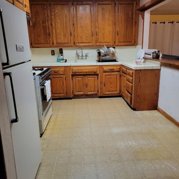 Hoadley Construction - Kitchen before - A kitchen with wooden upper and lower cabinets, a white refrigerator on the left, and a stove with a towel hanging on its handle. The countertop has dishes, a dish rack, and kitchen items. The floor is tiled with a beige pattern, and a ceiling light illuminates the space. The kitchen opens to another room through a pass-through window with a wooden frame. - General Contractors Near Me - Redondo Beach, California