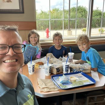 Culver's - Enjoying lunch! - A man and three children are seated at a table inside a restaurant with large windows showing greenery outside. They have fast food and drinks on the table. - Fast Food Near Me - Hoffman Estates, Illinois