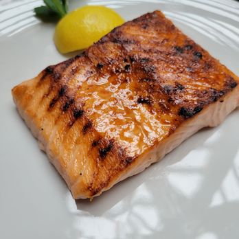 RH Chicago | The Gallery at the Three Arts Club - Salmon - A cooked salmon fillet with grill marks and a glaze, served with a lemon wedge on a white plate. - Furniture Stores Near Me - Chicago, Illinois