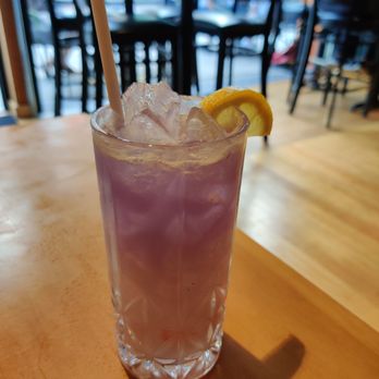 Claim 52 Kitchen - Queen cocktail - A purple-colored cocktail with ice in a tall glass, garnished with a lemon wedge and a straw, on a wooden table in a restaurant setting. - Gastropubs Near Me - Eugene, Oregon