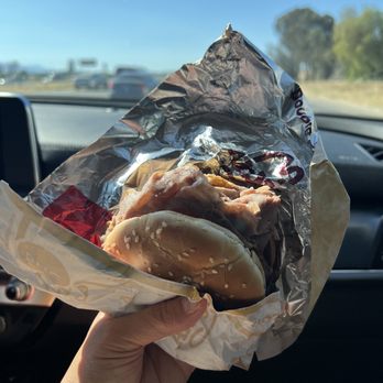 Arby's - a hand holding a sandwich wrapped in foil - A person is holding a burger wrapped in foil and paper inside a car, with the highway and blue sky visible through the windshield. The burger appears to contain meat and a sesame seed bun. The background shows moving vehicles and trees, indicating a road trip or drive-through meal. - Fast Food Near Me - Temecula, California