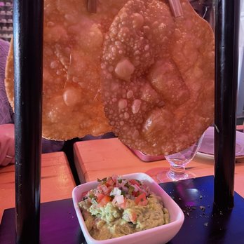 Travesias - Travesias Guacamole - Three large, crispy fried tortilla chips with bubbles are hanging on a stand, with a bowl of guacamole topped with diced tomatoes in front. - Latin American Near Me - Bronx, New York