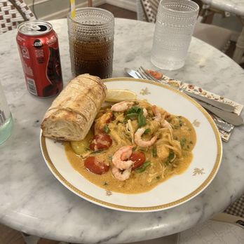 August Cafe - Spaghetti de mer - A marble table holds a plate of seafood pasta featuring shrimp, cherry tomatoes, and green herbs, with a bread roll and lemon wedge. Two drinks are present: a glass of soda with a yellow-striped straw and a glass of water. The setting appears to be a restaurant with patterned chairs and a neutral interior background. - Cafes Near Me - New York, New York