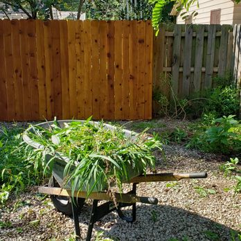 Sharp Edge Tree & Landscape - A garden scene featuring a wheelbarrow filled with greenery, enclosed by a wooden fence and a gravel pathway. - Landscaping Near Me - Mentor, Ohio