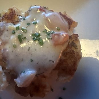 Becket's - Salmon cakes - A close-up image of a fried food item, possibly a potato or seafood dish, topped with a creamy white sauce and garnished with chopped green onions. The dish is presented on a white plate, with some sauce and herbs scattered around. The lighting is soft, highlighting the textures of the fried exterior and the sauce. - New American Near Me - Oshkosh, Wisconsin