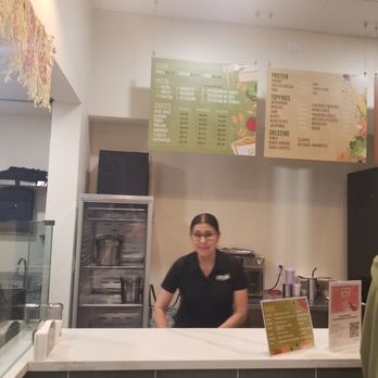 Pastissimo - A woman wearing glasses and a black shirt stands behind a white counter in a restaurant or cafe. Behind her, a menu is hanging on the wall, listing various food and drink options. The ceiling has bright lights, and there is a refrigerator and some kitchen equipment visible in the background. - Italian Near Me - Austin, Texas