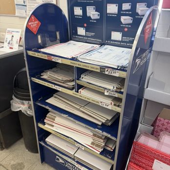 US Post Office - A blue USPS mailbox contains multiple shelves stocked with various mailing envelopes, including priority mail, flat rate, and express envelopes. The mailbox features signage on top with information about domestic delivery options and prices, and labels indicating the types of envelopes available. The mailbox is situated indoors, next to a trash can and other postal supplies. - Post Offices Near Me - Methuen, Massachusetts