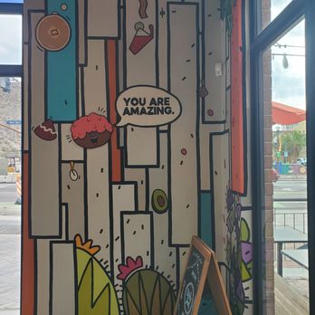 Daily Jam - Breakfast & Brunch Restaurant - Inside - The image shows a colorful, abstract mural on an interior wall of a cafe, with various geometric shapes, a smiling character with pink hair, and a speech bubble that reads 'You are amazing.' Natural light enters through large windows, revealing a street scene outside. A small chalkboard sign is positioned in front of the mural, and the overall setting suggests a casual, artistic atmosphere. - New American Near Me - Tempe, Arizona