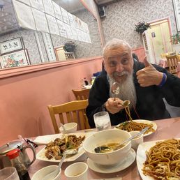 Loved the menus on the mirror in both English and Mandarin! - A man with a long white beard and black jacket is seated at a table in a Chinese-Korean restaurant, giving a thumbs-up and holding a spoonful of noodles.