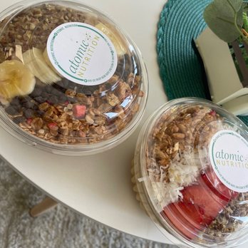 Atomic Nutrition - Tropical and monkey bowl - Two clear plastic containers of granola bowls from Atomic Nutrition are positioned on a white table. The bowls contain granola with toppings such as banana slices, shredded coconut, and strawberries. One container has a label with the Atomic Nutrition logo. A green woven placemat and a potted plant are also visible on the table, along with a textured floor beneath. - Juice Bars & Smoothies Near Me - Elgin, Texas