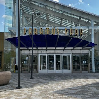 Paramus Park - The image shows the main entrance of Paramus Park shopping mall, featuring glass doors and a blue canopy above the entrance. The building has a modern design with glass and metal elements, and the signage displays the mall's name in gold letters. The foreground includes a paved walkway with a large planter and street lamps, and the sky above is partly cloudy with sunlight illuminating the scene. - Shopping Centers Near Me - Paramus, New Jersey