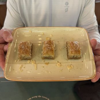 Moda Cafe and Restaurant - Baklava - A person presents a plate with three pieces of pistachio baklava at Moda Cafe and Restaurant, a Turkish pastry shop. - Turkish Near Me - Chantilly, Virginia