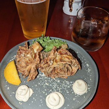 CODE RED - Artichoke and IPA - A round gray plate contains fried food garnished with a lemon wedge, some greens, and three dollops of mayonnaise. Two beverages are on the table: a tall glass of beer with foam and a shorter glass of soda with ice. The setting appears to be in a restaurant or bar with a dark wooden table and a napkin holder in the background. - Cocktail Bars Near Me - Washington, DC, District of Columbia