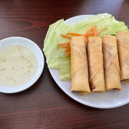 Spring Rolls - A plate with four Vietnamese spring rolls, accompanied by lettuce, shredded carrots, and a small bowl of dipping sauce, on a dark wooden table.