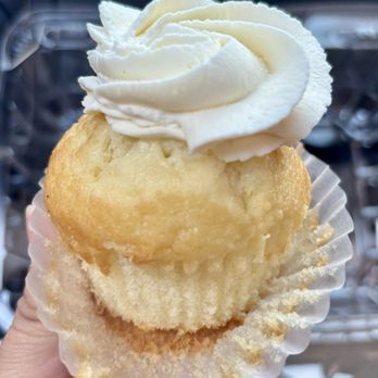Brooklyn Cupcake - Dulce de Leche Cupcake - A cupcake with vanilla frosting swirled on top and a cookie placed above the frosting, held in a white paper liner. The background is blurred, showing a metallic surface and possibly a display or storage area. - Cupcakes Near Me - Brooklyn, New York